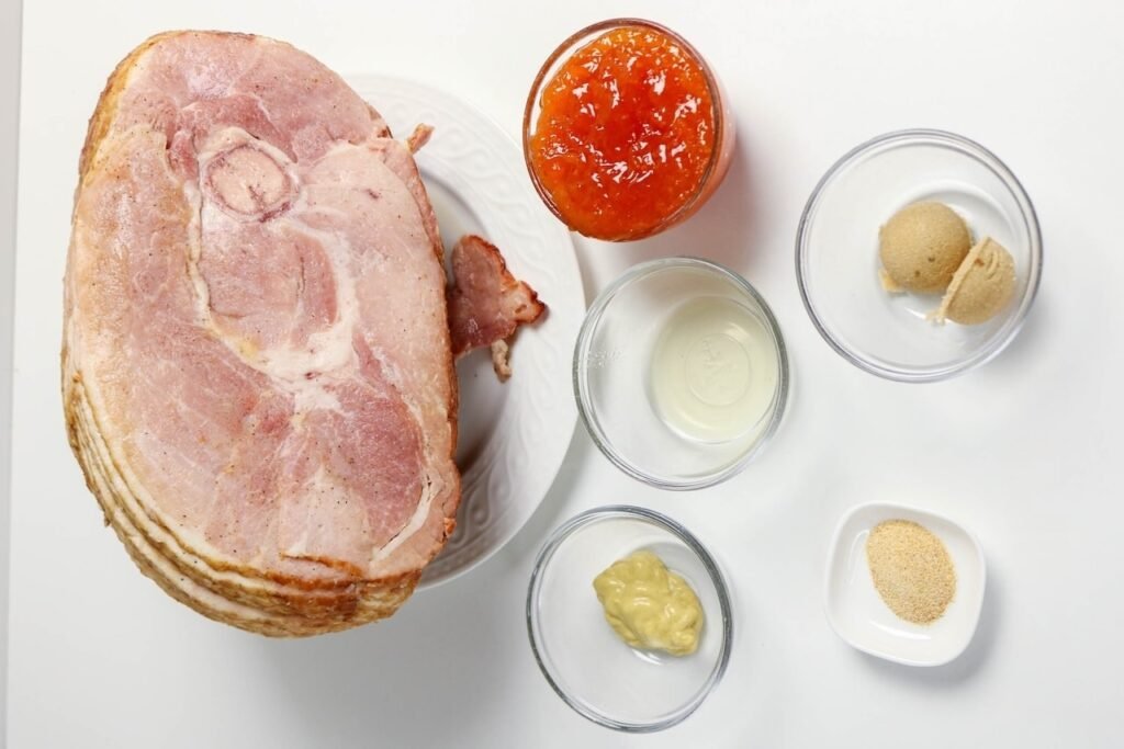 Ingredients for apricot glazed ham including spiral-cut ham, apricot preserves, Dijon mustard, brown sugar, apple cider vinegar, and garlic powder arranged on a white surface.
