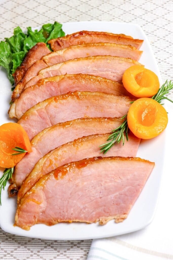 Close-up of meat on a white platter, topped with golden caramelized glaze and garnished with fresh rosemary and apricot halves.