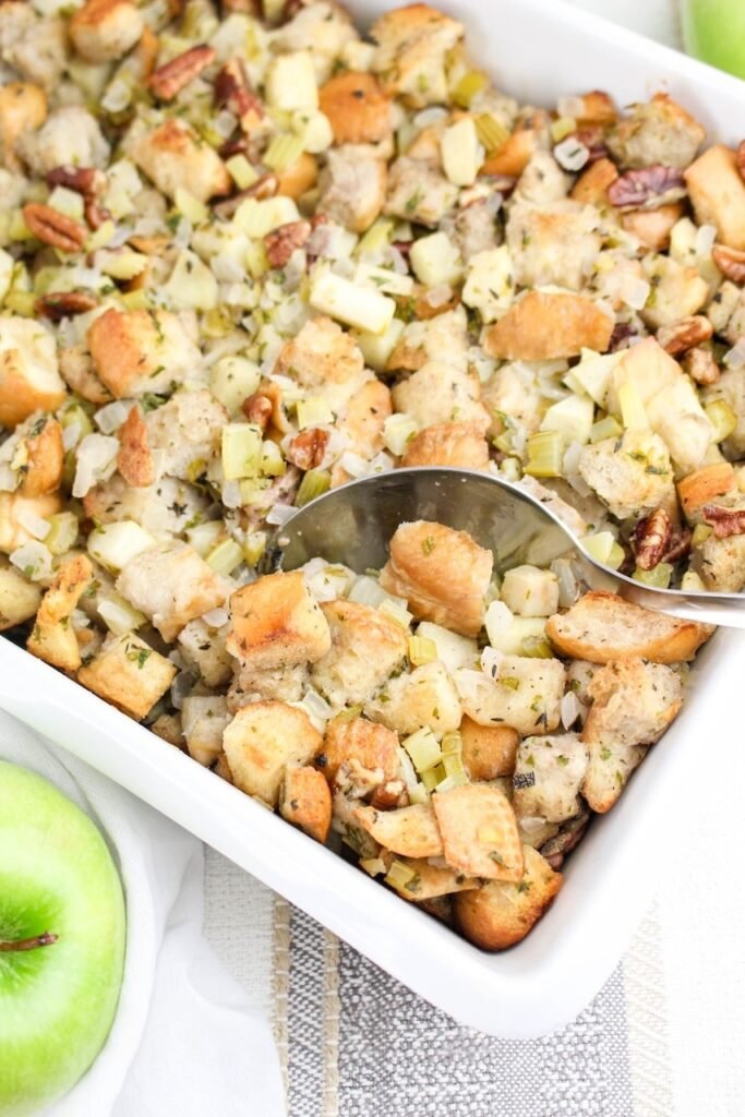 Golden baked herb and apple stuffing with bread cubes, apples, celery, and pecans in a white casserole dish with a serving spoon.