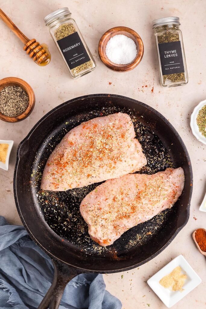 Raw breast seasoned with herbs, salt, pepper, and paprika in a cast-iron skillet, ready to sear for oven recipe.