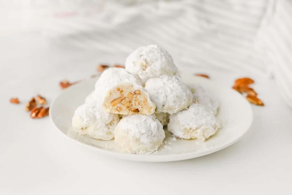Stack of buttery pecan snowball cookies coated in powdered sugar on a white plate, with one cookie cut open to show the chopped pecan filling.