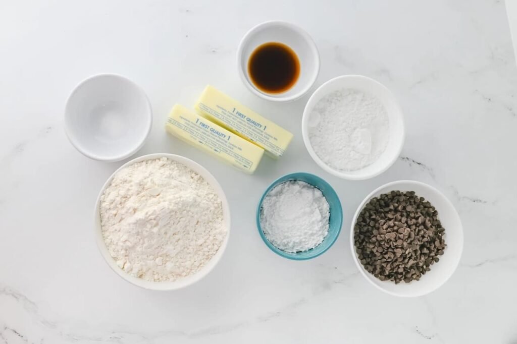 Ingredients for chocolate chip snowball cookies arranged on a white marble surface including butter, flour, powdered sugar, vanilla, and mini chocolate chips.