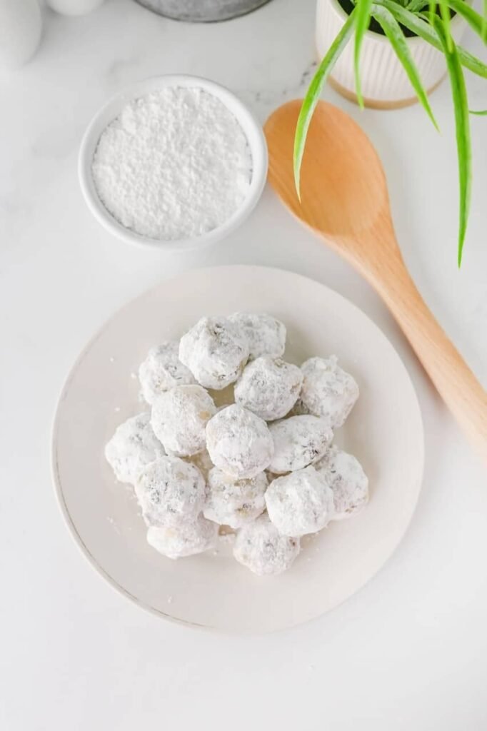 Overhead view of chocolate chip snowball cookies coated in powdered sugar on a white plate beside a bowl of sugar and wooden spoon.