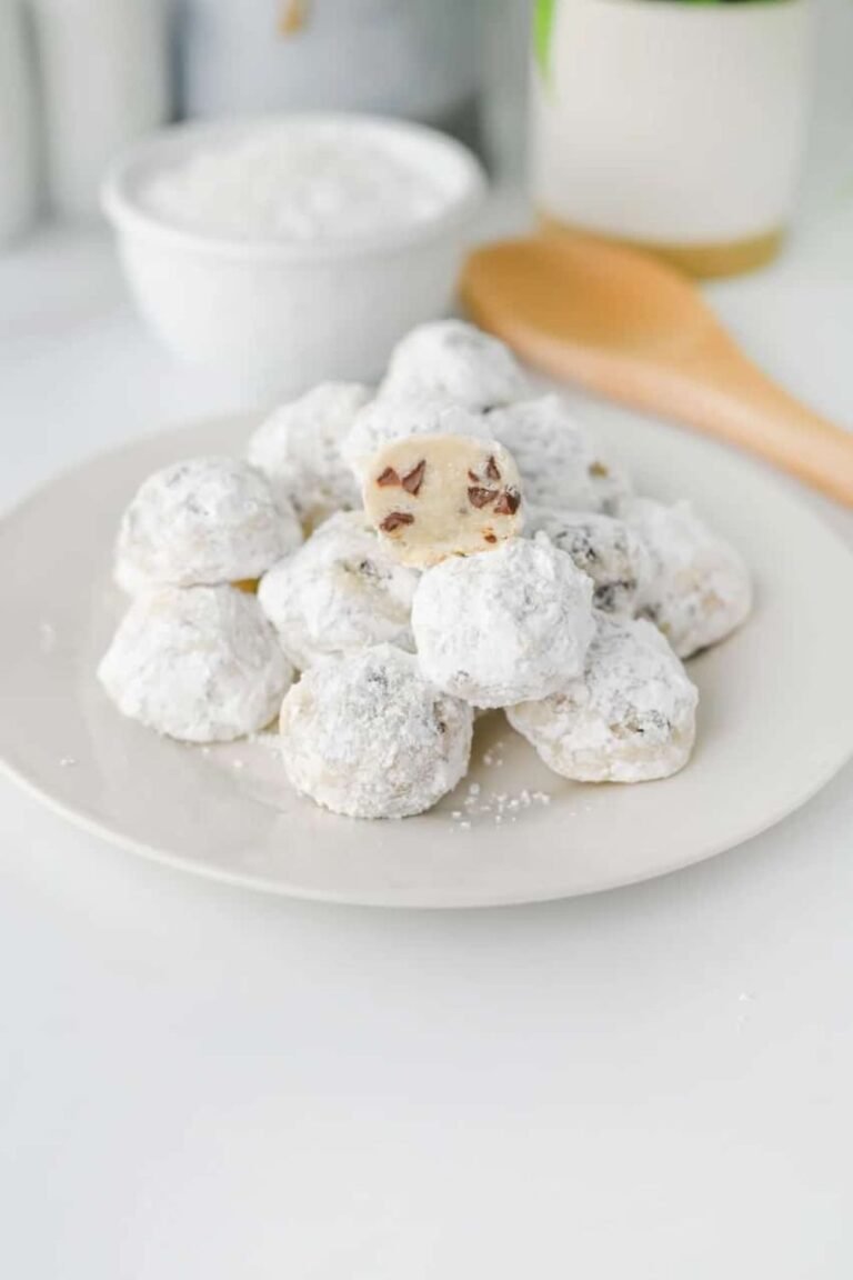 Plate of chocolate chip snowball cookies coated in powdered sugar with one cookie cut open to show mini chocolate chips inside.
