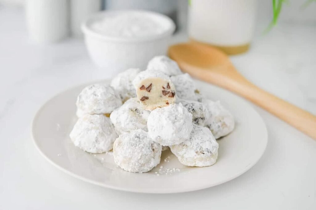 Plate of buttery chocolate chip snowball cookies coated in powdered sugar with one cookie showing chocolate chips inside.