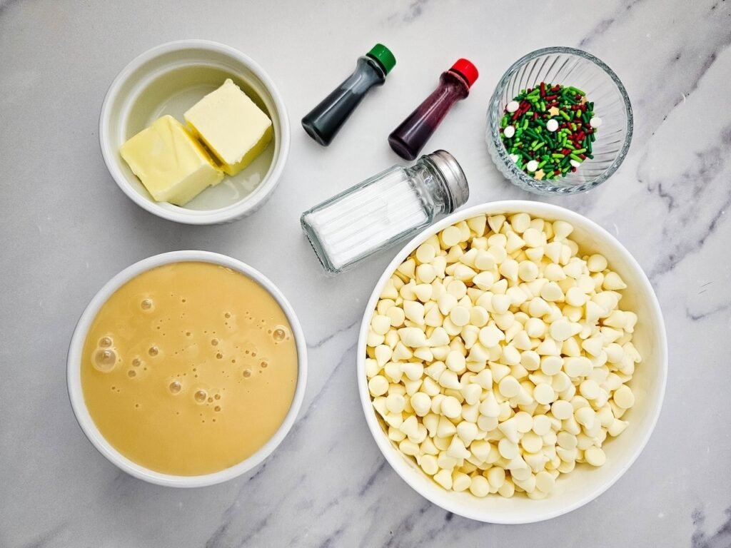 Ingredients for easy Christmas fudge recipe including white chocolate chips, sweetened condensed milk, butter, salt, food coloring, and holiday sprinkles on marble counter.
