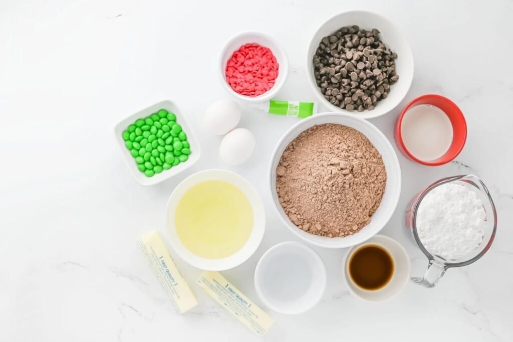 Overhead view of Grinch Brownie ingredients including cocoa mix, butter, eggs, chocolate chips, neon green food coloring, red heart sprinkles, and green M&Ms arranged on a white surface.