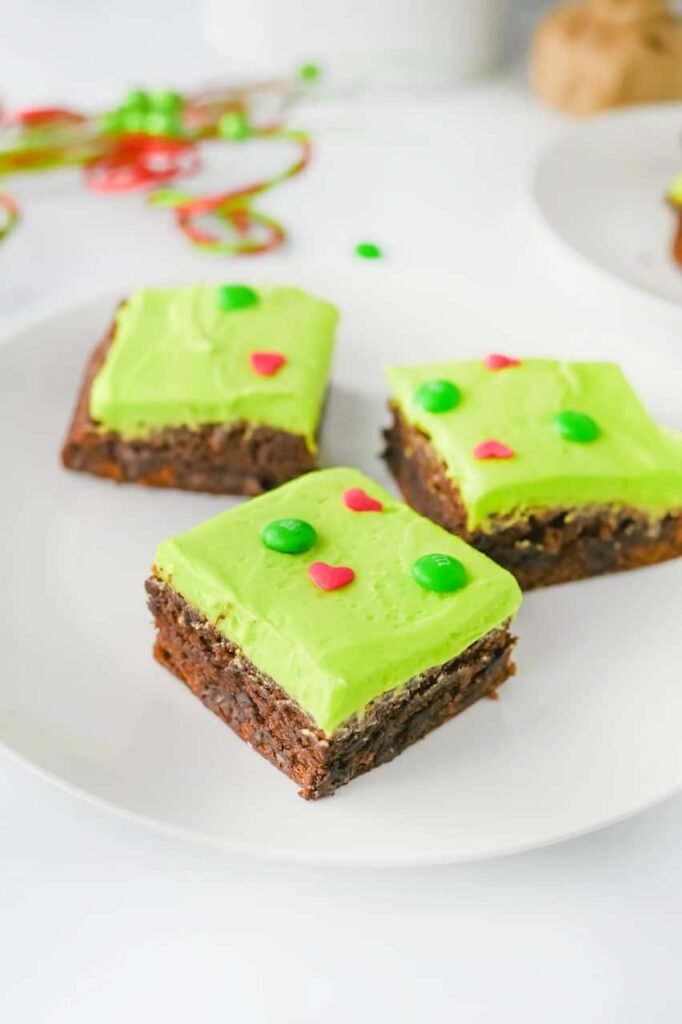 Three Grinch Brownies topped with green frosting, red heart sprinkles, and green M&Ms served on a white plate for a festive holiday dessert.