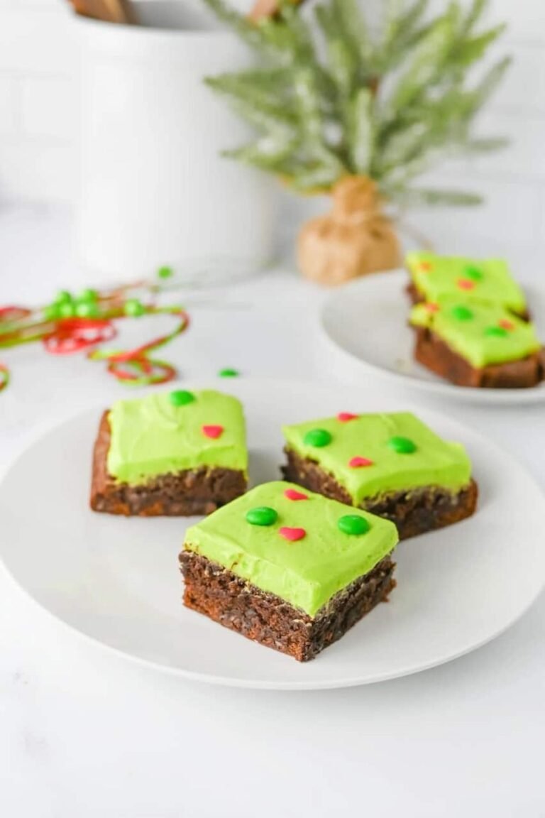 Three Grinch Brownies topped with bright green frosting, red heart sprinkles, and green M&Ms served on a white plate with festive holiday decor in the background.