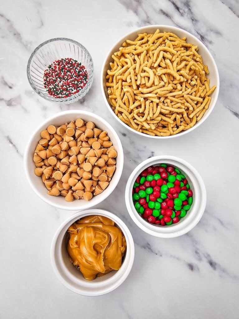 ingredients including chow mein noodles, butterscotch chips, creamy peanut butter, red and green M&Ms, and festive sprinkles on a marble surface.