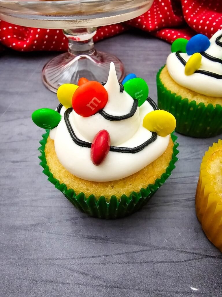Close-up of a Christmas Light Cupcake decorated with buttercream frosting, black icing, and colorful M&Ms designed to look like holiday lights.