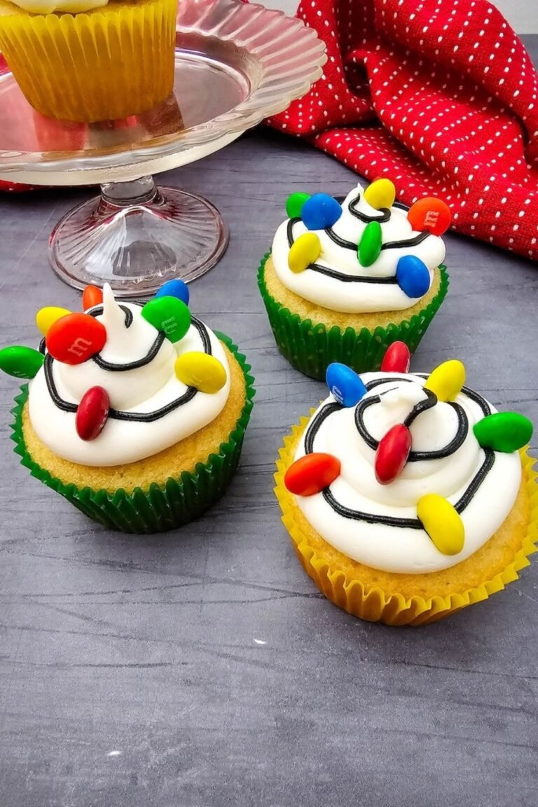 Featured image of Christmas Light Cupcakes topped with buttercream frosting, black icing, and colorful M&Ms arranged like festive holiday lights.
