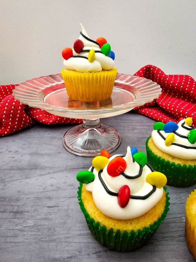 Christmas Light Cupcakes displayed on a glass stand, topped with buttercream frosting, black icing, and colorful M&Ms to look like festive holiday lights.
