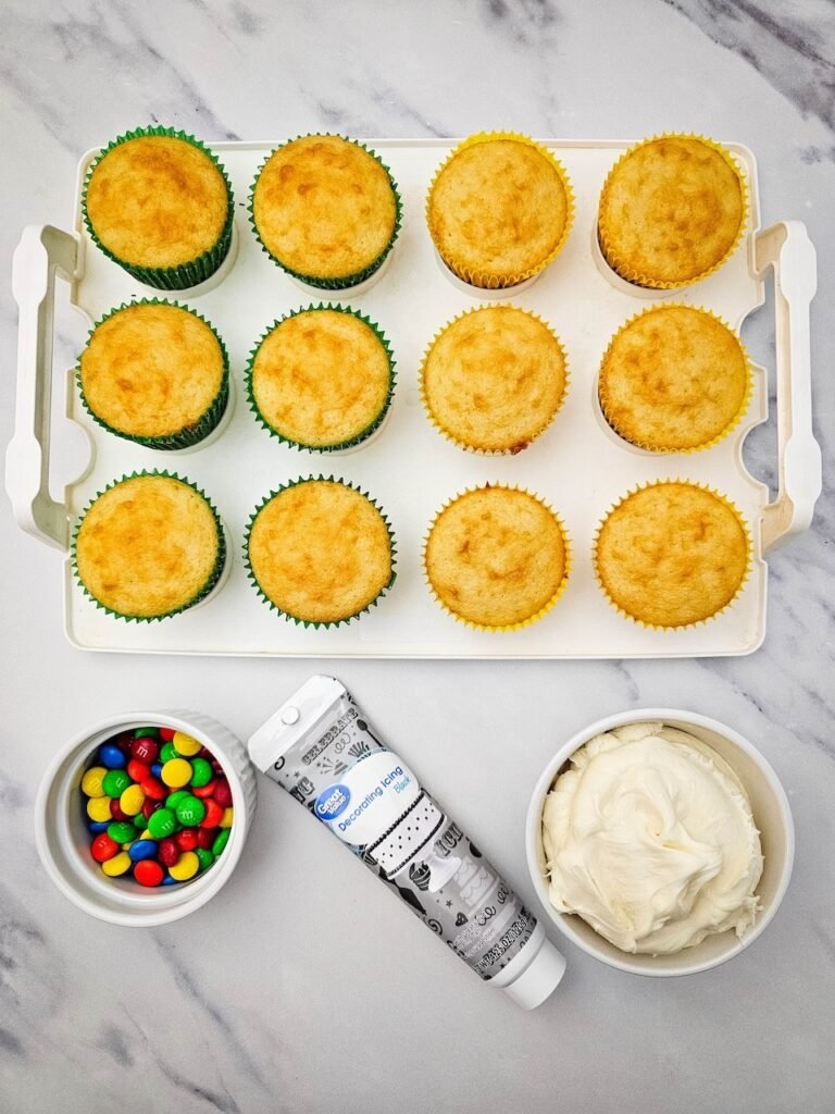 Ingredients for Christmas Light Cupcakes including vanilla cupcakes, buttercream frosting, black decorating gel, and colorful M&Ms on a white marble surface.