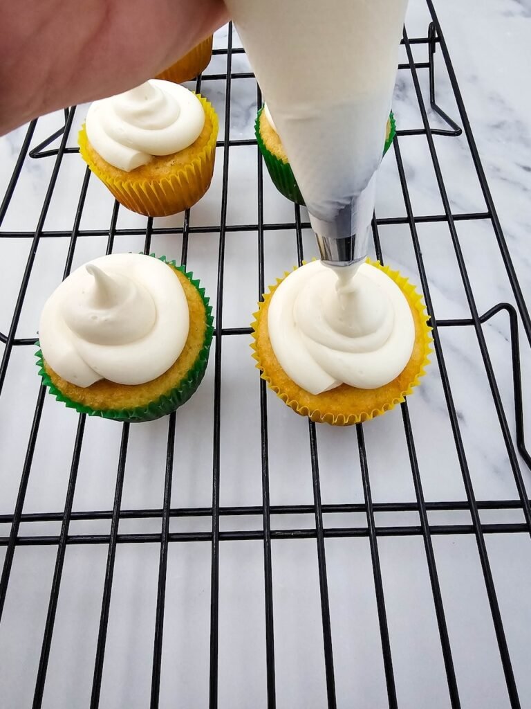 Piping smooth buttercream frosting onto vanilla cupcakes with yellow and green liners.