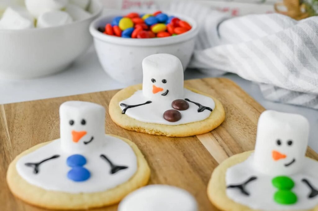 Melted snowman cookies decorated with marshmallow heads, icing faces, and colorful candy buttons on a wooden board with bowls of toppings in the background.