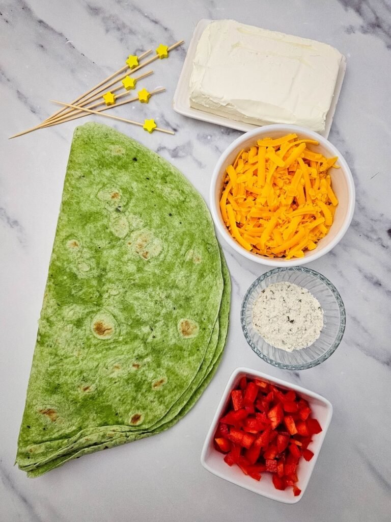 ingredients for Christmas tree pinwheels arranged on a marble surface. Fresh spinach tortillas are shown with a block of cream cheese, shredded cheddar cheese in a white bowl, diced red bell pepper, a small dish of ranch seasoning, and yellow star-shaped food picks for decorating the holiday appetizer.
