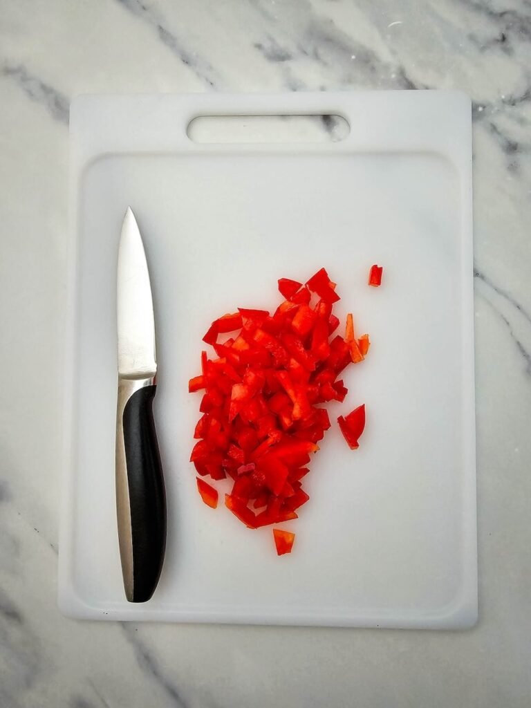 Finely diced red bell pepper on a white cutting board with a paring knife. This ingredient adds color and flavor to Christmas pinwheels, making the holiday appetizer look festive when rolled into tortillas with cream cheese and cheese.
