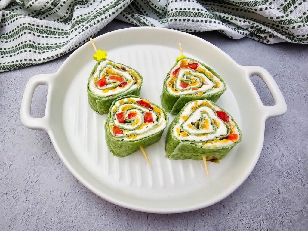 Four Christmas pinwheels displayed on a round white platter with handles. Each pinwheel is made with spinach tortillas rolled with cream cheese, shredded cheddar, and diced red peppers, secured with skewers topped with yellow star picks. A green striped towel in the background highlights the holiday appetizer presentation.