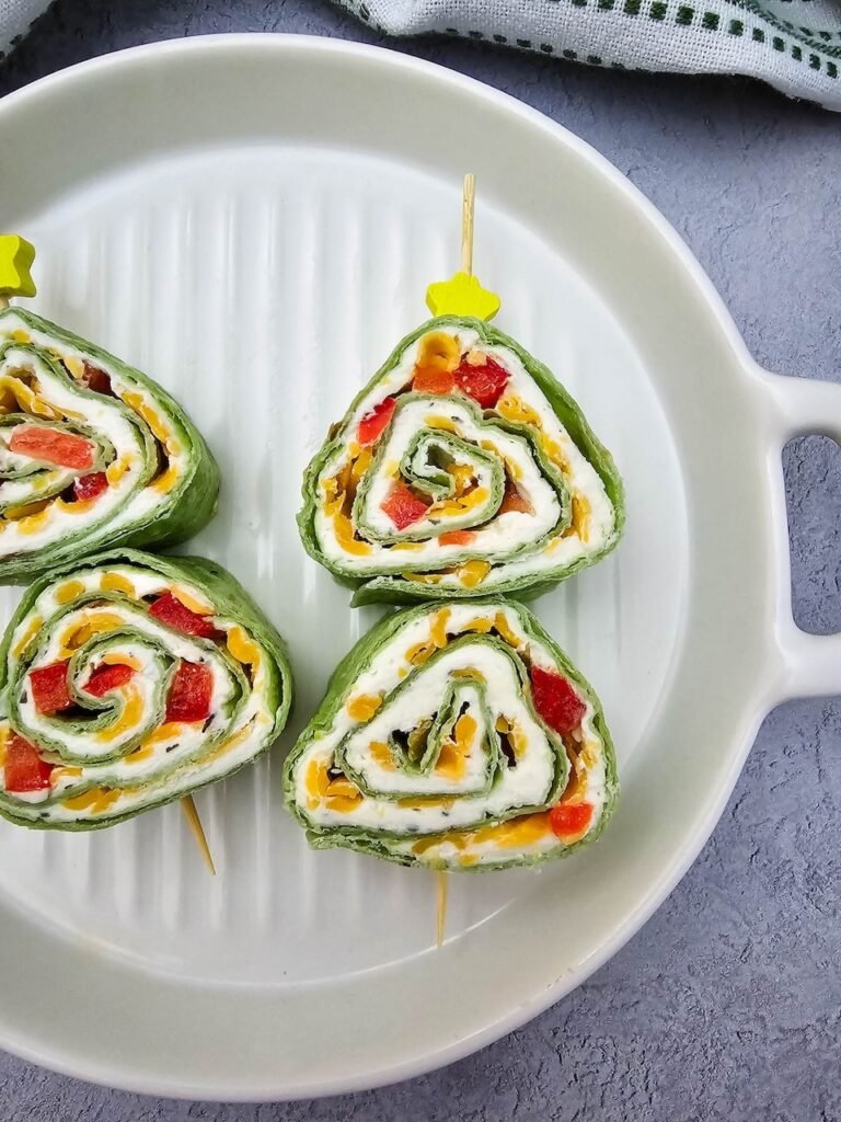 Close-up of Christmas tree pinwheels arranged on a white platter. The pinwheels are made with spinach tortillas rolled with cream cheese, ranch seasoning, shredded cheddar cheese, and finely diced red peppers. Each slice is triangular and held with a food pick topped with a yellow star, creating a festive holiday appetizer look.