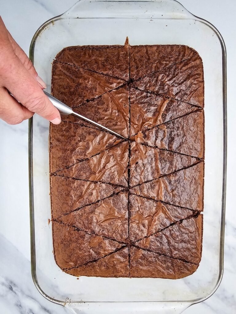 A hand cuts a cooled 9x13-inch pan of brownies into triangle shapes using a knife in a glass baking dish. The brownies are sliced into evenly sized wedges that will be decorated with red and white frosting to create festive Santa Hat Brownies for a Christmas dessert tray.