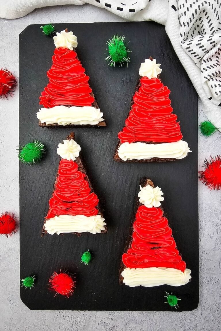 Four Santa Hat Brownies decorated with red and white frosting on a black slate board surrounded by green and red holiday pom-poms.