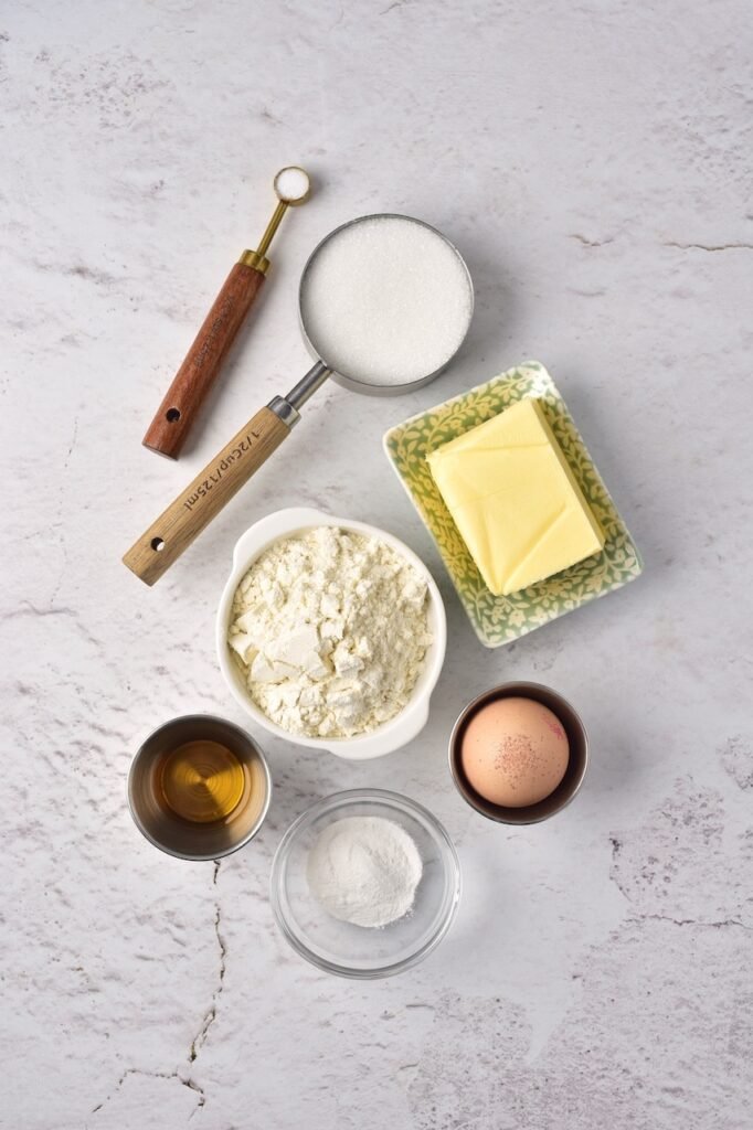 Overhead view of ingredients including butter, flour, sugar, egg, vanilla extract, and baking powder on a light marble surface.