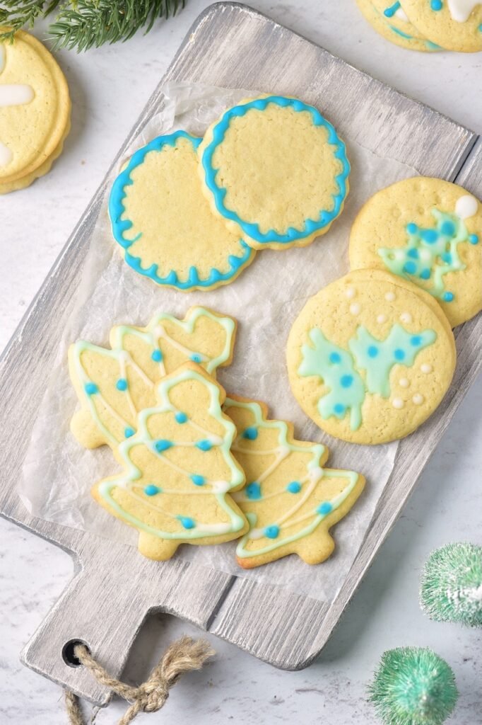 Decorated Christmas sugar cookies displayed on a rustic wooden serving board, including Christmas tree-shaped cookies and round cut-out cookies decorated with blue, green, and white glaze. The baked cookies have a soft, buttery texture and are arranged for holiday serving and decorating inspiration.