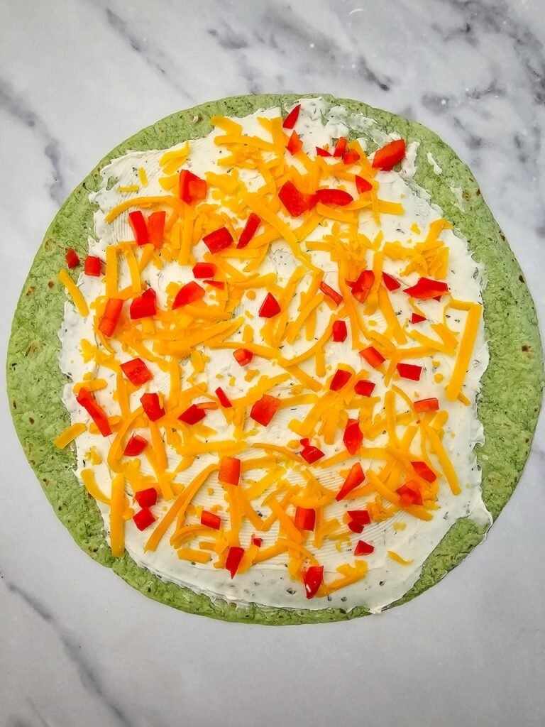 Spinach tortilla spread with cream cheese and ranch seasoning, topped with shredded cheddar cheese and finely diced red bell peppers. This step shows how to layer ingredients before rolling into a log to create the festive holiday appetizer.