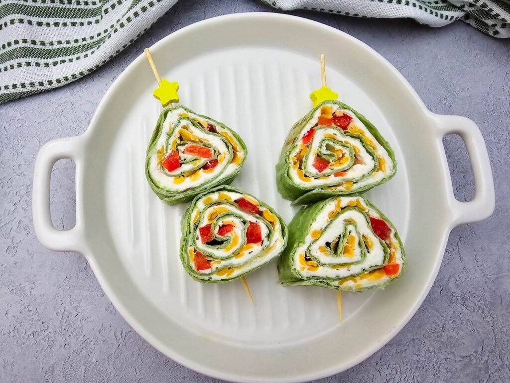 Christmas tree pinwheels made with spinach tortillas, cream cheese, shredded cheese, and red pepper arranged as holiday appetizer roll-ups.