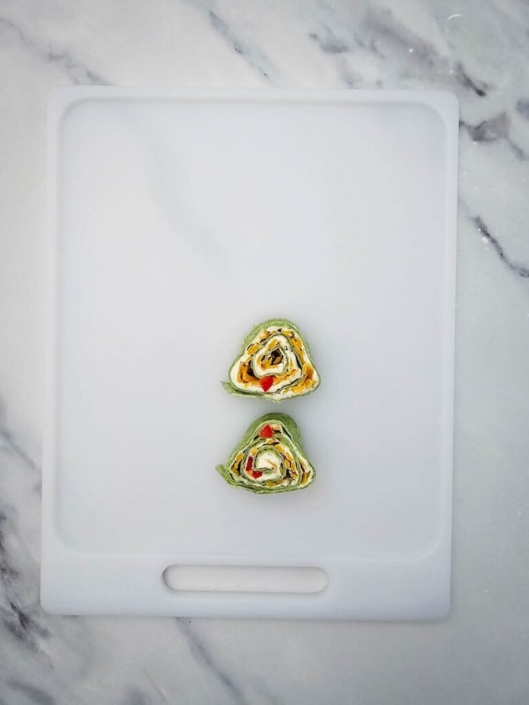 two slices of Christmas tree pinwheels placed on a white cutting board, starting to form the triangular tree shape. Each pinwheel is made with spinach tortillas filled with cream cheese, shredded cheddar cheese, and diced red peppers, arranged step by step into a festive holiday appetizer tree.