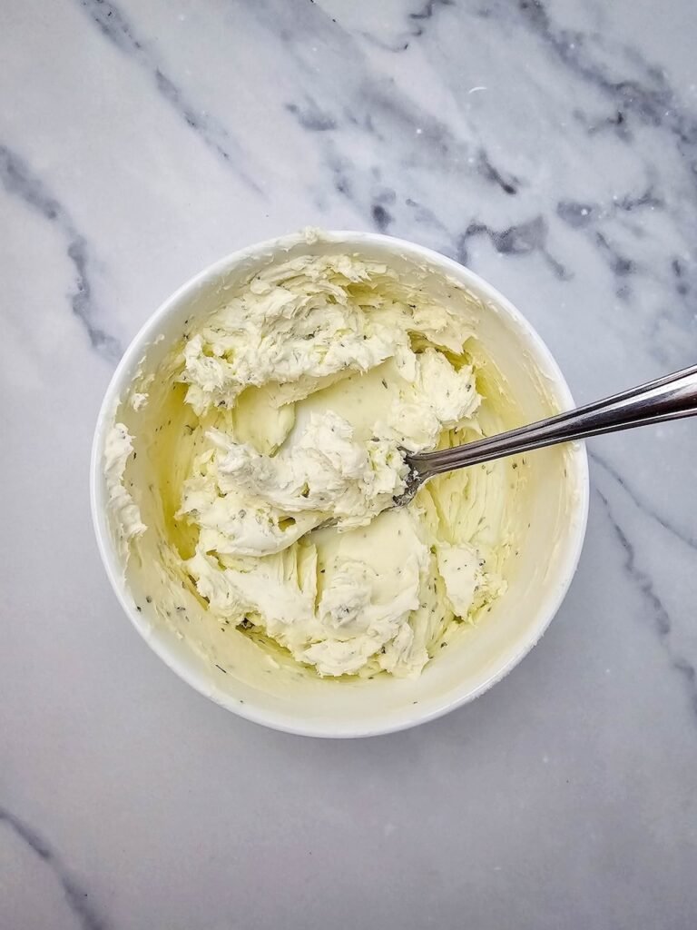Bowl of softened cream cheese mixed with ranch seasoning and a spoon on a marble surface. This creamy filling is the base for Christmas tree pinwheels, spread onto tortillas before adding shredded cheese and red pepper for the festive holiday appetizer.