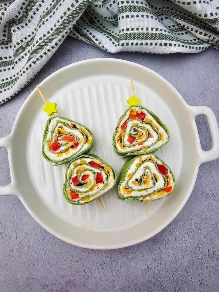 four Christmas tree pinwheels arranged on a round white platter. Each pinwheel is made with spinach tortillas rolled with cream cheese, shredded cheddar cheese, and red peppers, then secured with skewers topped with yellow star picks. Served as a festive holiday appetizer with a green striped towel in the background.