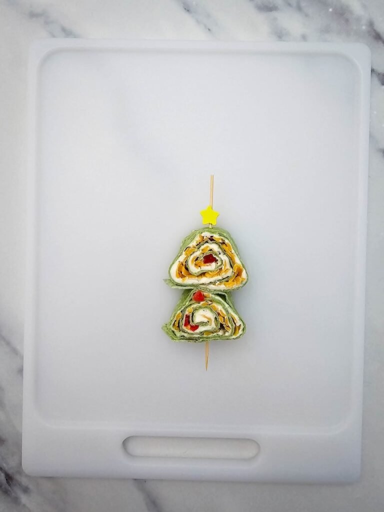 two slices of Christmas tree pinwheels stacked vertically on a skewer with a yellow star food pick on top, creating the look of a mini holiday tree. Made with spinach tortillas, cream cheese, shredded cheddar, and red peppers, this appetizer step shows how to decorate pinwheels for Christmas.