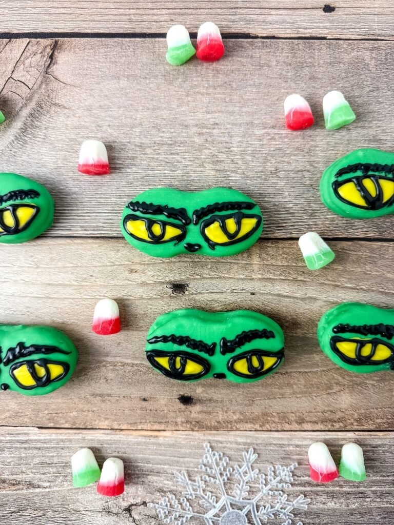 Close-up of Grinch Nutter Butter cookies with yellow eyes and black eyebrows surrounded by red and green holiday candies.
