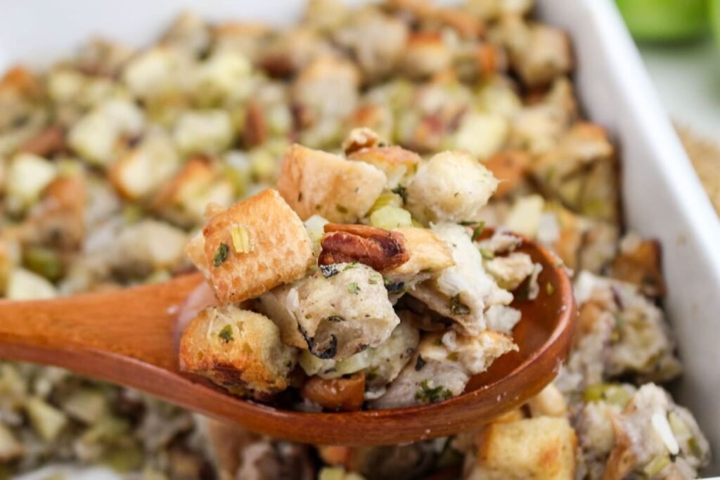 Close-up of baked herb and apple stuffing on a wooden spoon, showing golden bread cubes, apples, celery, and pecans.