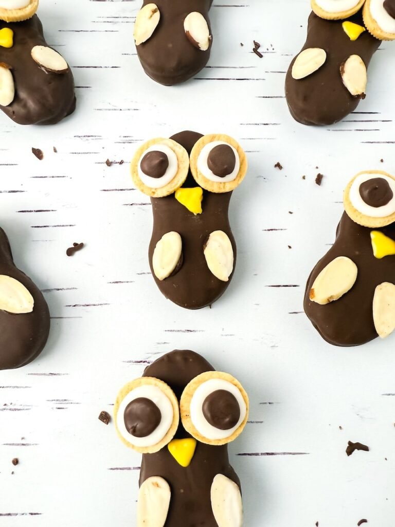 Close-up view of Nutter Butter owl cookies on a white birch-patterned surface, each coated in chocolate and decorated with cookie eyes, yellow beaks, and almond slice wings for a cute fall dessert idea.