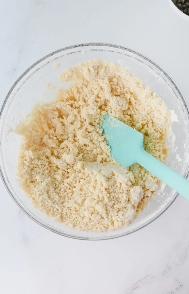 Mixing butter mixture with flour to form crumbly dough, using a blue spatula in a glass bowl.