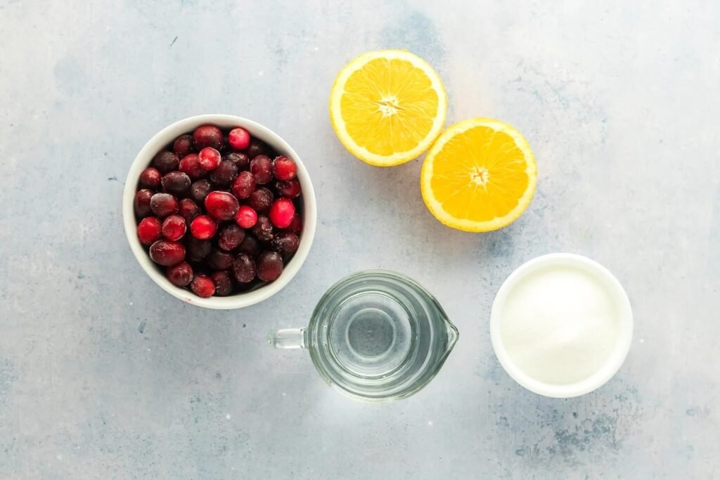 ingredients for homemade cranberry sauce laid out on a light surface, including fresh cranberries, halved oranges, sugar, and water.