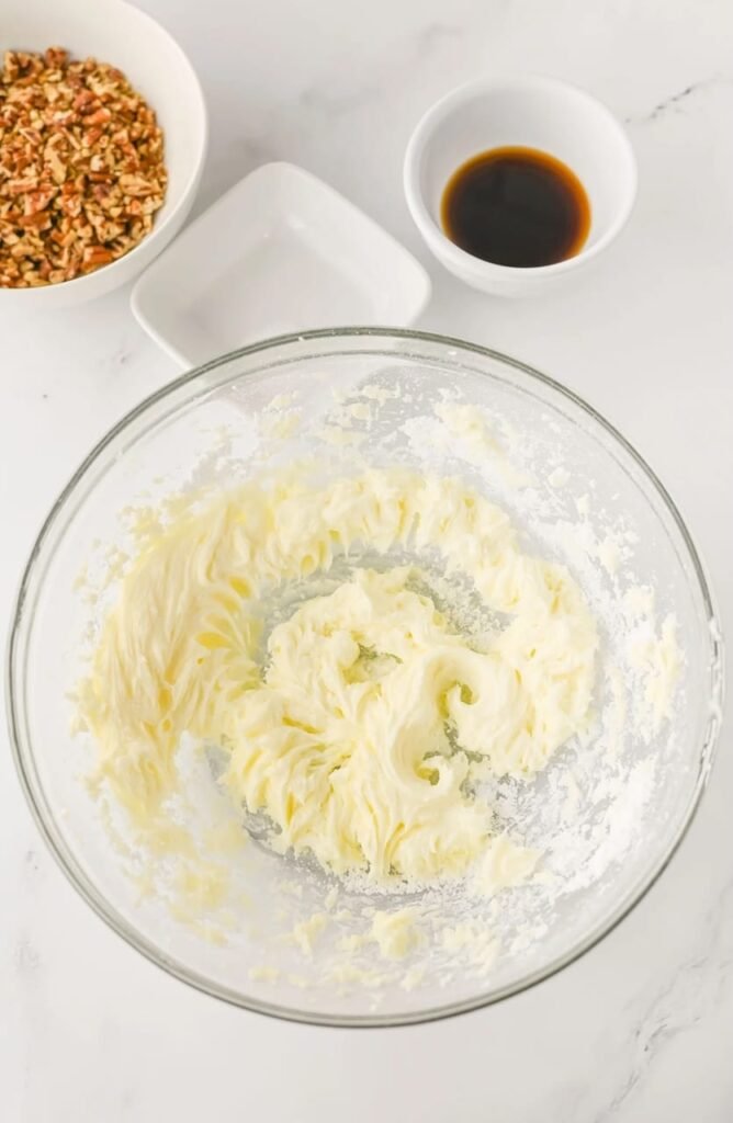 Butter and powdered sugar creamed together in a glass bowl with vanilla extract and chopped pecans nearby.