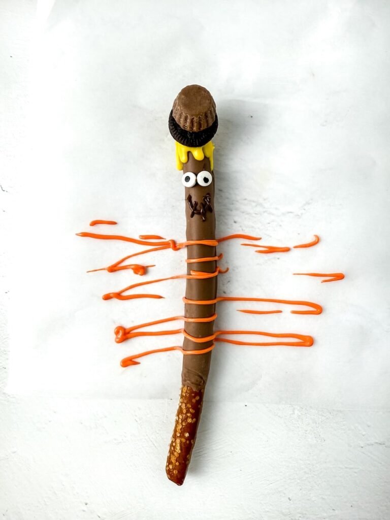 Finished rod decorated with candy eyes, yellow candy melt “straw” hair, a mini Oreo and peanut butter cup hat, and orange candy drizzle forming the scarf. The decorated pretzel rests on parchment paper, showing the final decorating step before serving this cute fall dessert.