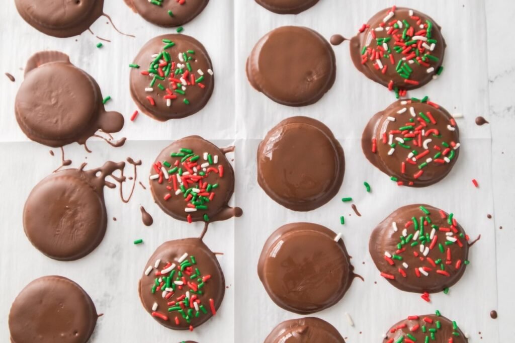 Chocolate-coated Ritz cracker thin mint cookies on parchment paper, half plain and half topped with red, green, and white sprinkles, showing the decorating step for no-bake Christmas Thin Mint Cookies.