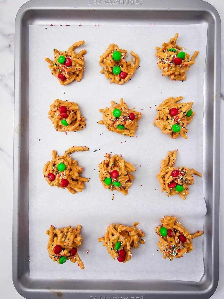 Christmas haystack cookies topped with red and green M&Ms and festive sprinkles on a parchment-lined baking sheet before setting.