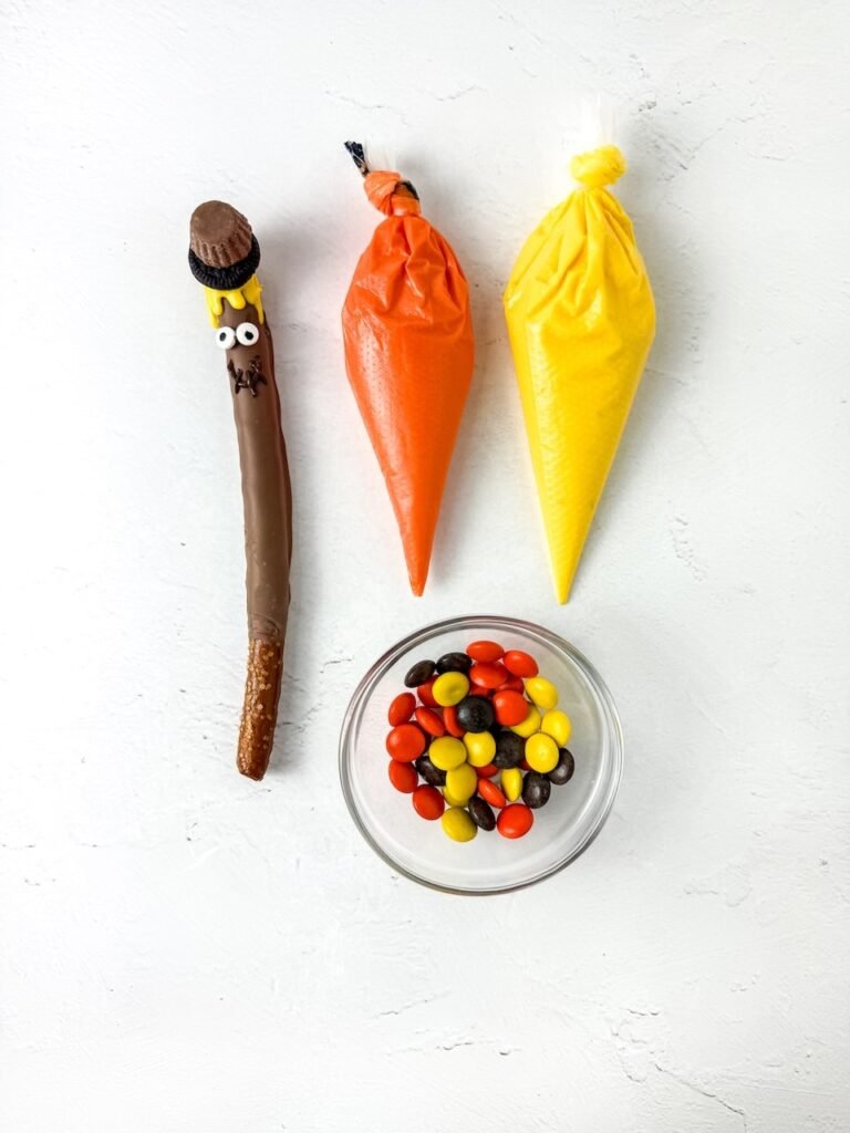 Step five decorating showing a chocolate-coated pretzel with candy eyes, yellow candy melt hair, and an Oreo and peanut butter cup hat. Orange and yellow piping bags filled with melted candy and a bowl of Reese’s Pieces are arranged on a white background for decorating the fall treats.