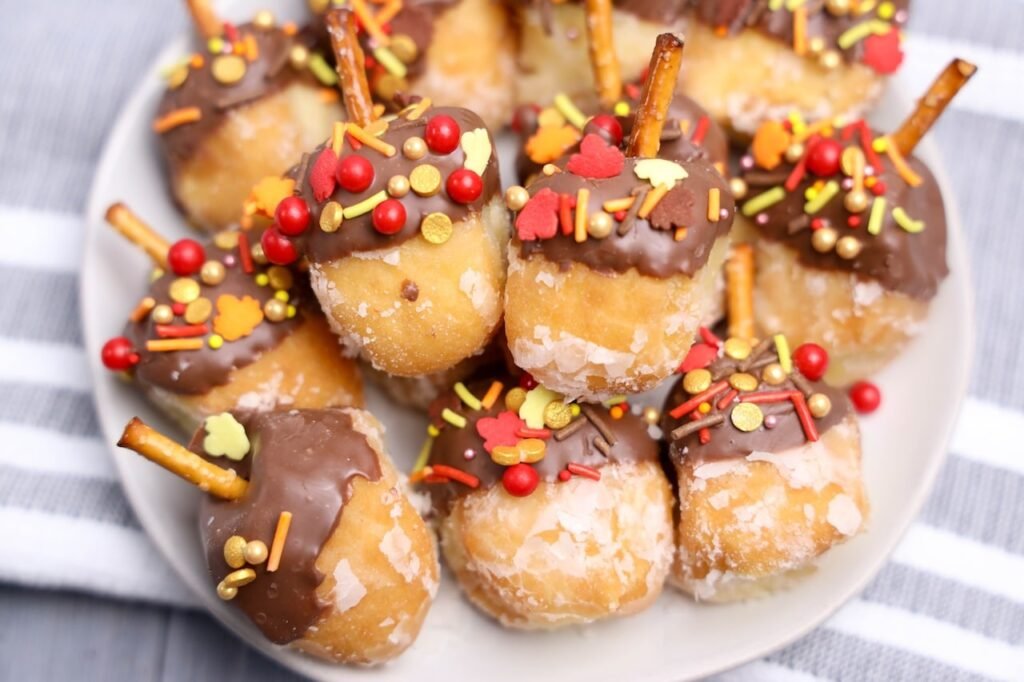 Easy acorn donut holes dipped in chocolate and topped with fall sprinkles and pretzel sticks on a white plate.