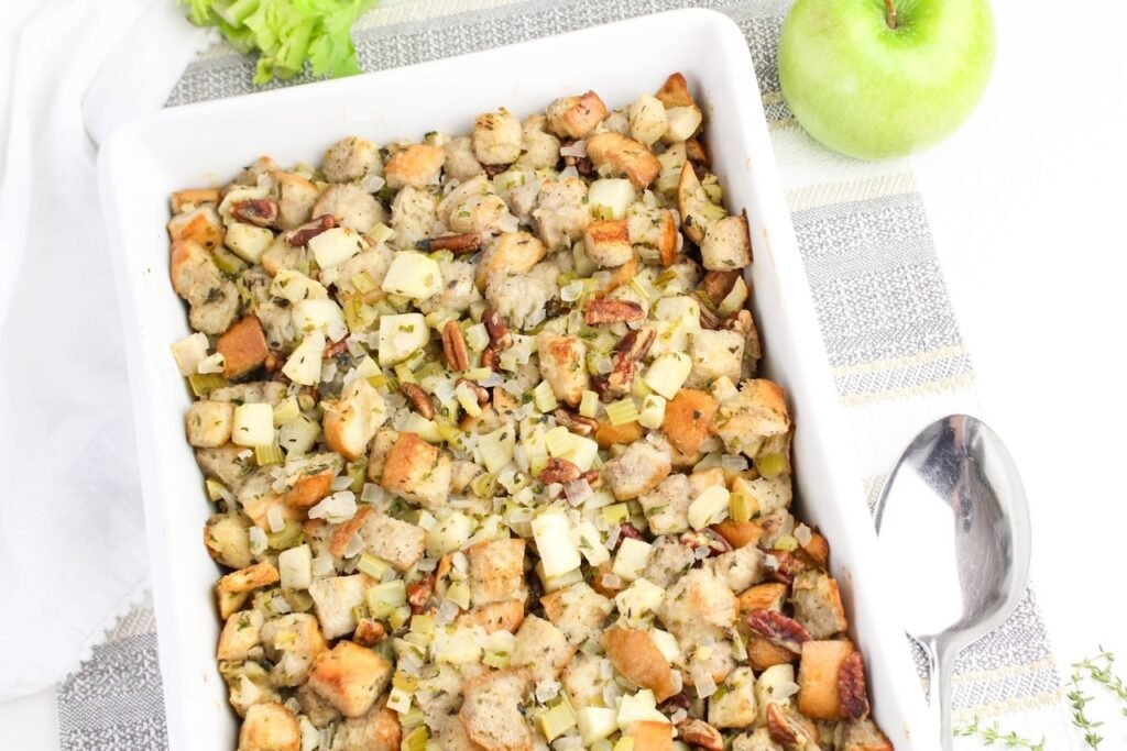Baked herb and apple stuffing in a white casserole dish with golden bread cubes, apples, celery, and pecans, styled with fresh apples on the side.