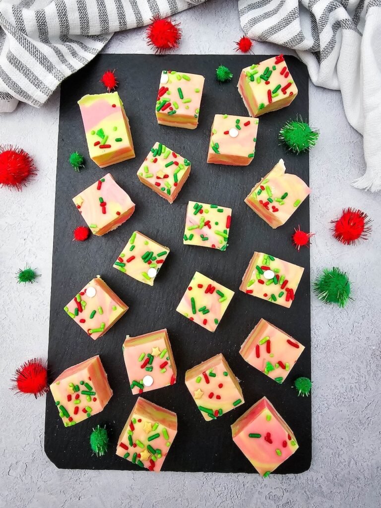 Overhead view of creamy swirled Christmas fudge squares topped with festive red and green sprinkles on a black slate board.
