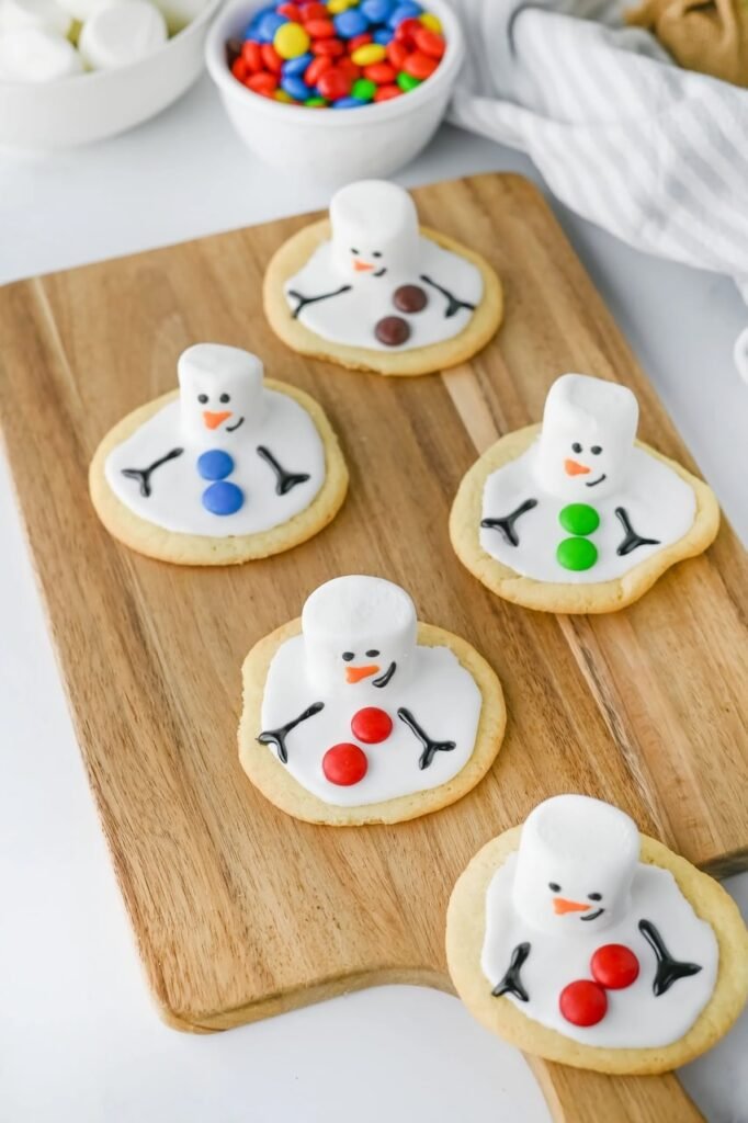 Finished melted snowman cookies decorated with marshmallow heads, icing arms, and colorful candy buttons on a wooden board.