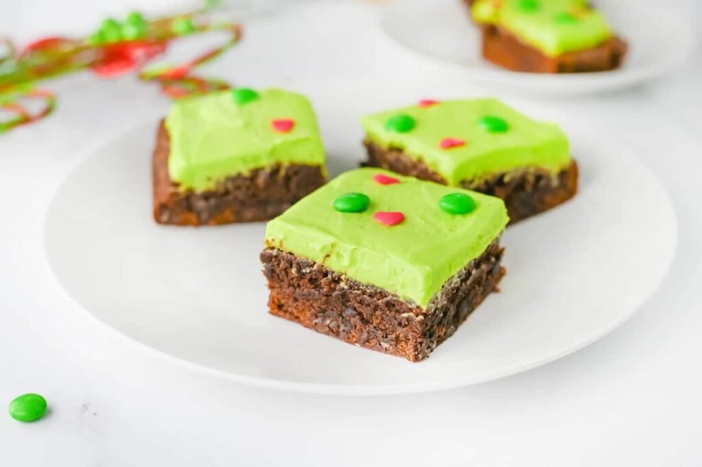 Fudgy Grinch Brownies topped with bright green frosting, red heart sprinkles, and green M&Ms sliced on a white plate for a festive Christmas dessert.