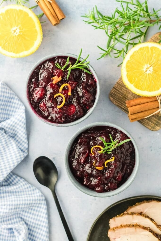 Two bowls of homemade cranberry sauce garnished with orange zest and rosemary, styled with cinnamon sticks and fresh orange halves.